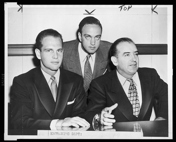 three men in suits are sitting at a table and one man is pointing to something