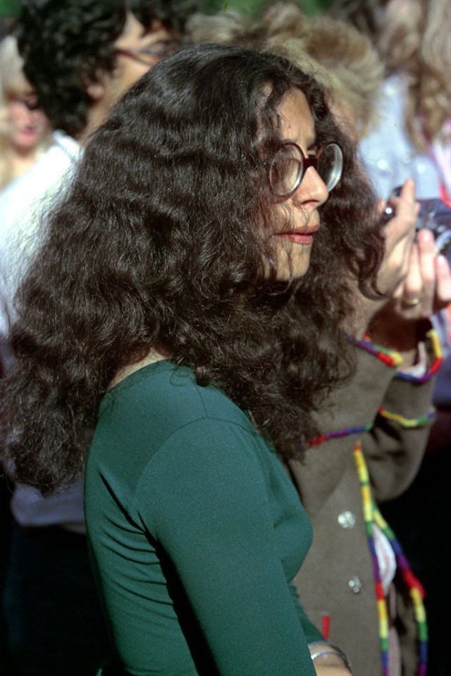 a woman with long curly hair and glasses standing in front of a group of people