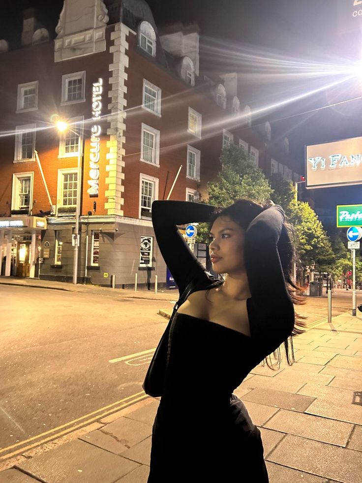 a woman in a black dress standing on the side walk at night with her hands behind her head