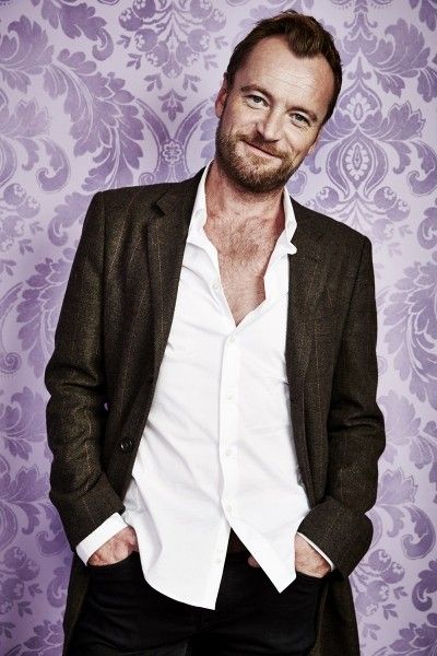 a man standing in front of a purple wall wearing a suit and white shirt with his hands in his pockets