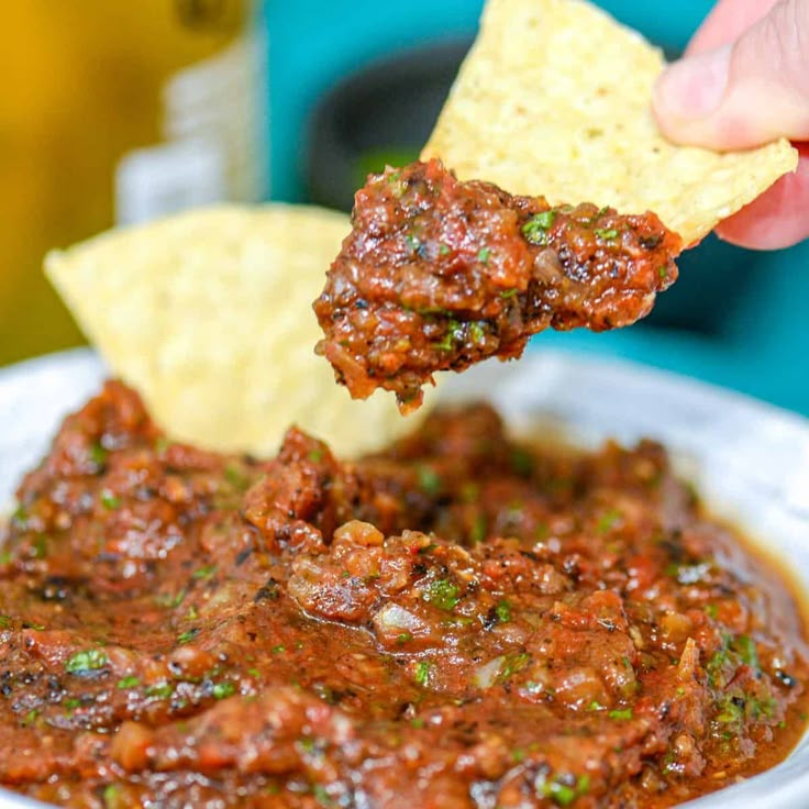 a hand holding a tortilla chip and dipping it into a bowl of salsa