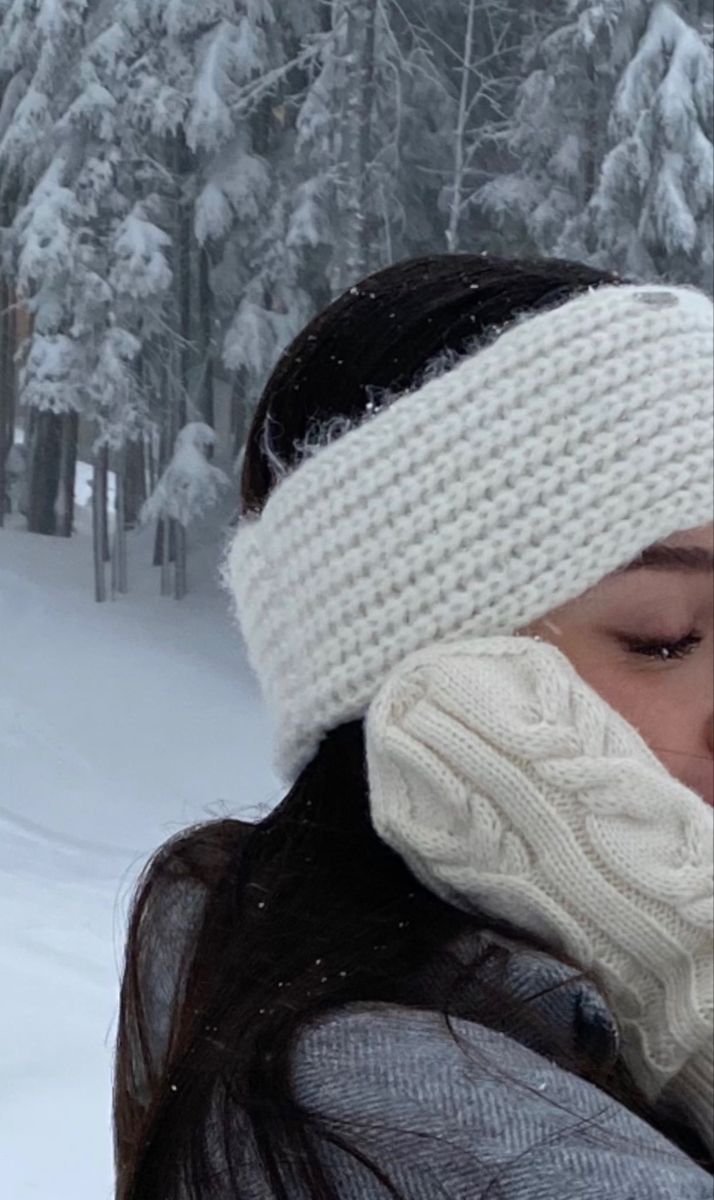 a woman wearing a white knitted hat and scarf in front of snow covered trees