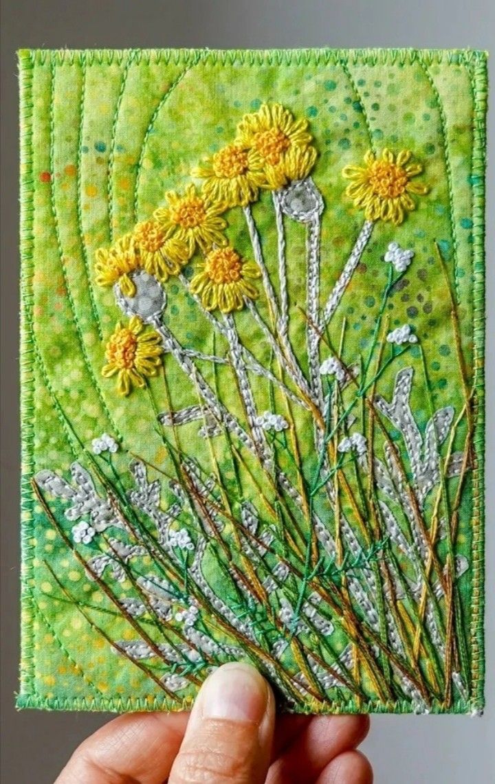 a hand holding up a piece of art with yellow flowers on green fabric and white thread