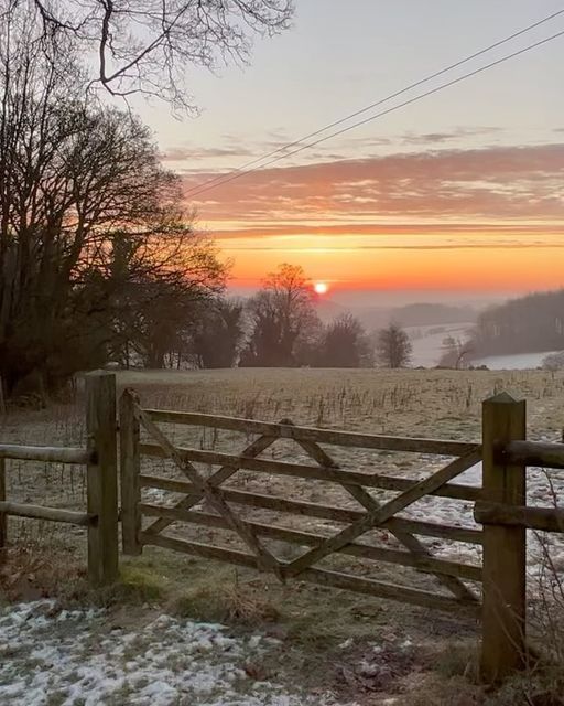 the sun is setting over a snowy field with a fence and trees in the foreground