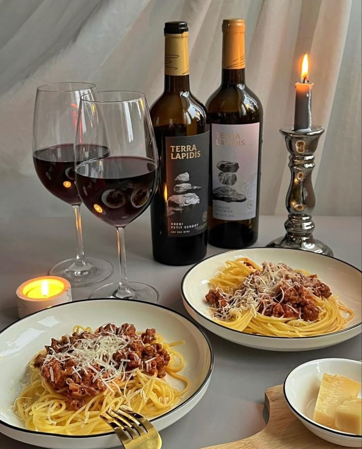 two plates of spaghetti and wine on a table with candles in front of the bottles