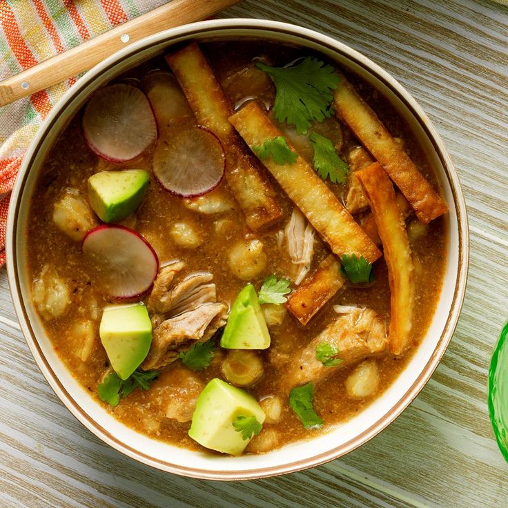 a bowl of soup with meat, potatoes and avocado on a wooden table