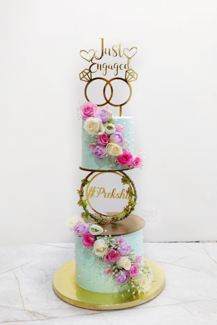 a three tiered cake with flowers on top and the words just engaged spelled in gold