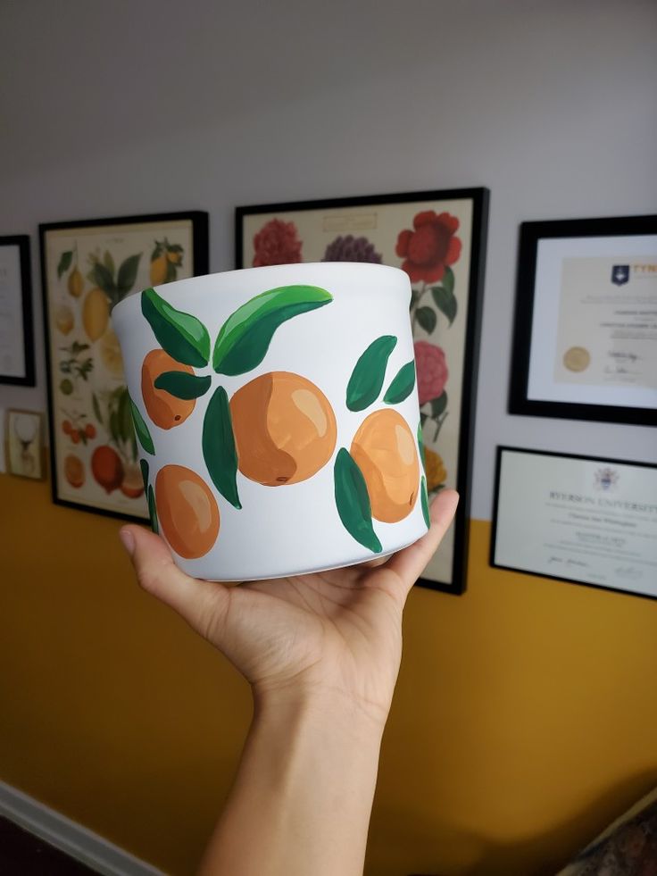 a person holding up a cup with oranges painted on it in front of some framed pictures