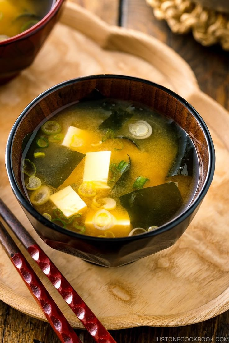 a bowl of soup with tofu and other vegetables on a wooden plate next to chopsticks