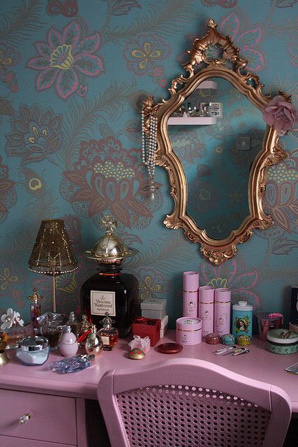 a pink desk with a mirror, lamp and other items on top of it in front of a floral wallpaper