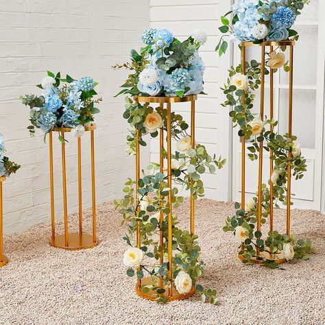 three gold vases with blue and white flowers in them on carpeted area next to brick wall