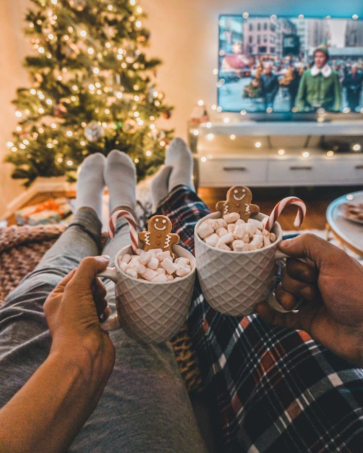 two people holding cups filled with marshmallows in front of a christmas tree