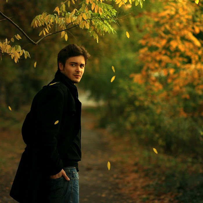 a young man standing in front of a tree with leaves falling from it's branches