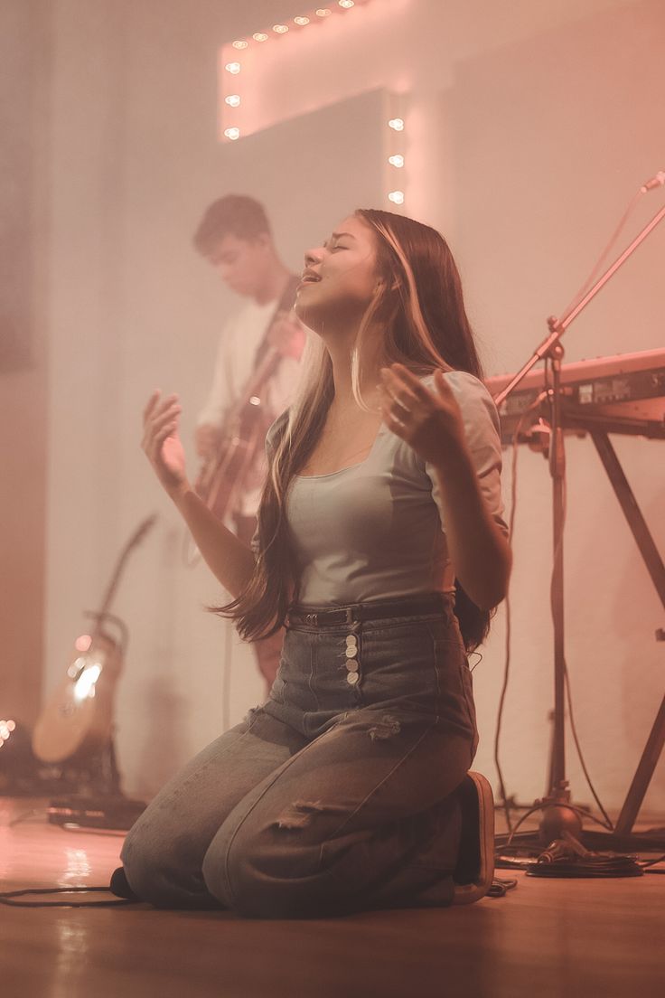 a woman sitting on the floor singing into a microphone while another man stands behind her