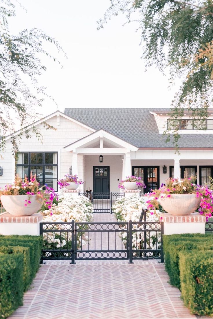 a white house with pink and purple flowers in the front yard, surrounded by hedges