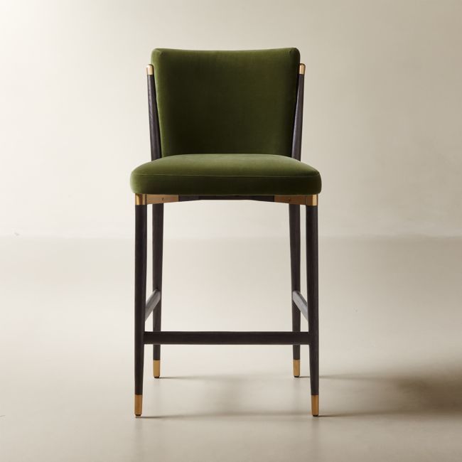 a green velvet bar stool with gold trim around the armrests and back rests against a white wall