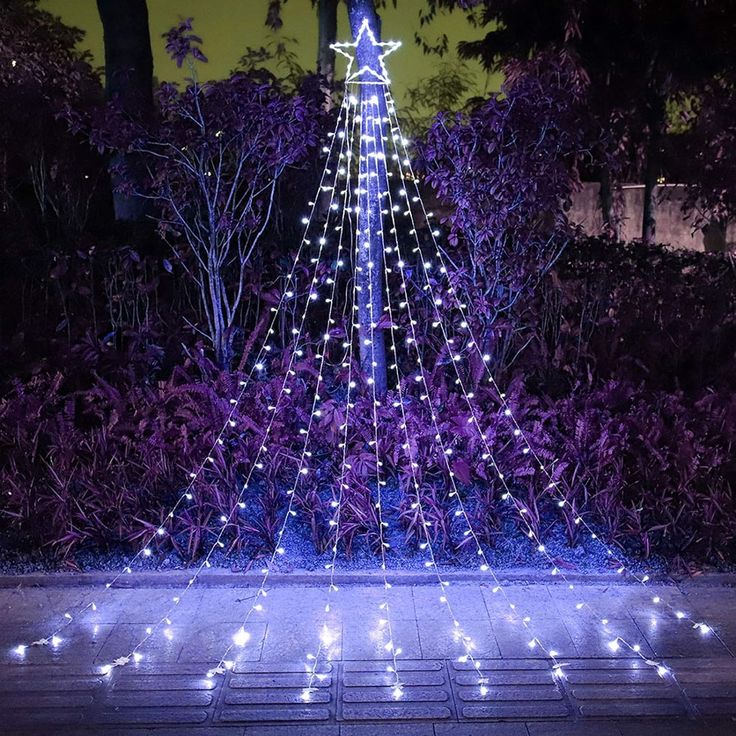 a lighted christmas tree in the middle of a garden with purple bushes and trees around it