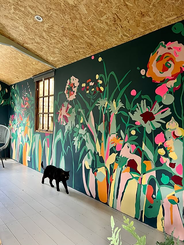 a black cat is walking in front of a colorful wall with flowers and plants on it