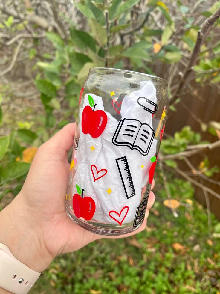 a hand holding up a glass with an apple and book design on the bottom, in front of some trees