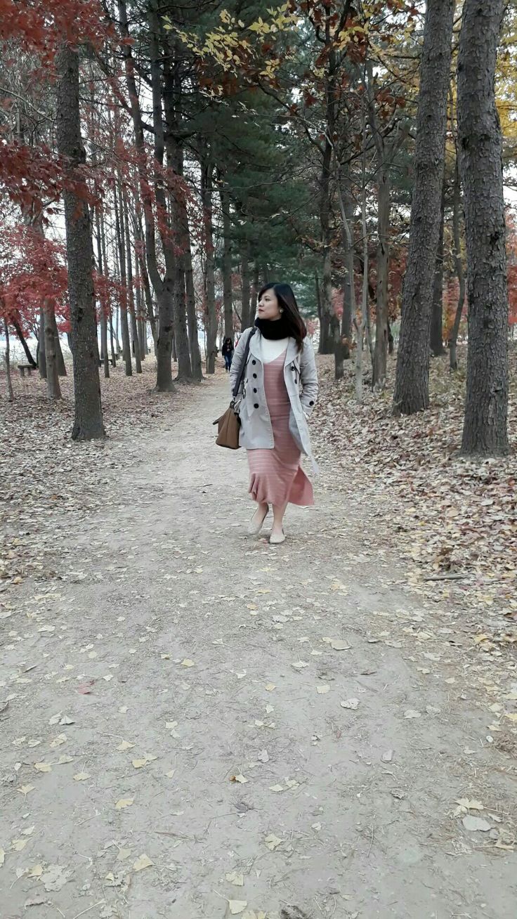 a woman walking down a dirt road in the woods with trees and leaves all around her