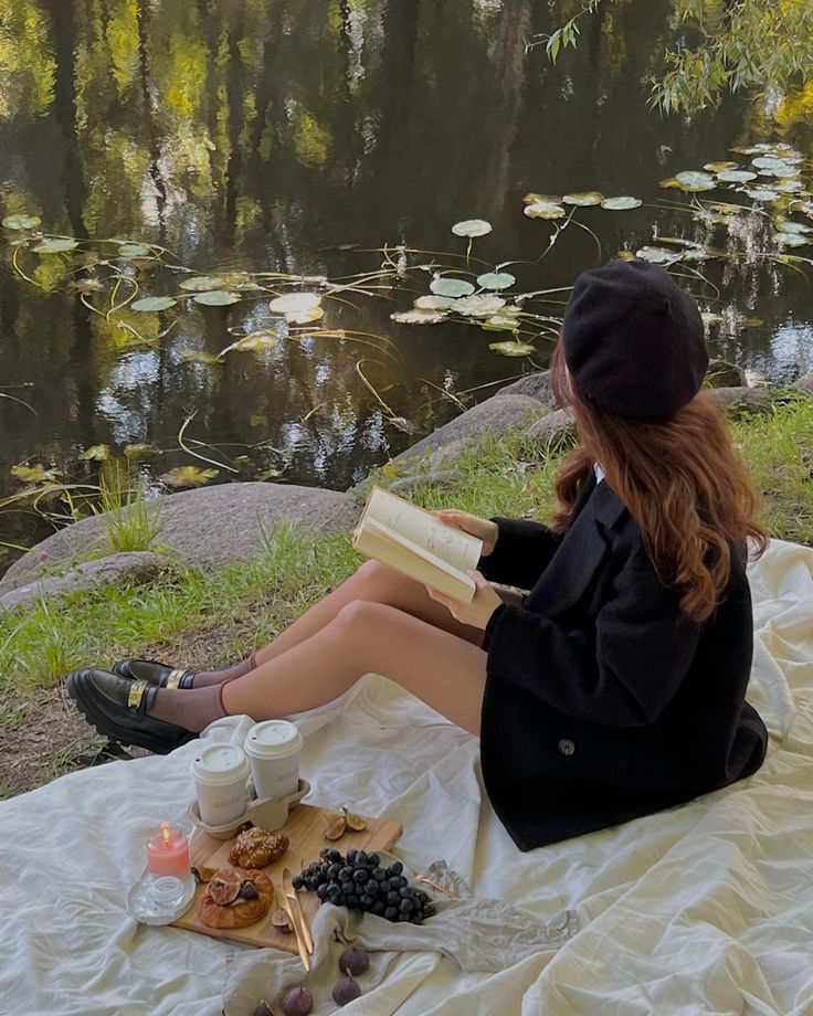 a woman sitting on a blanket next to a pond reading a book and eating food