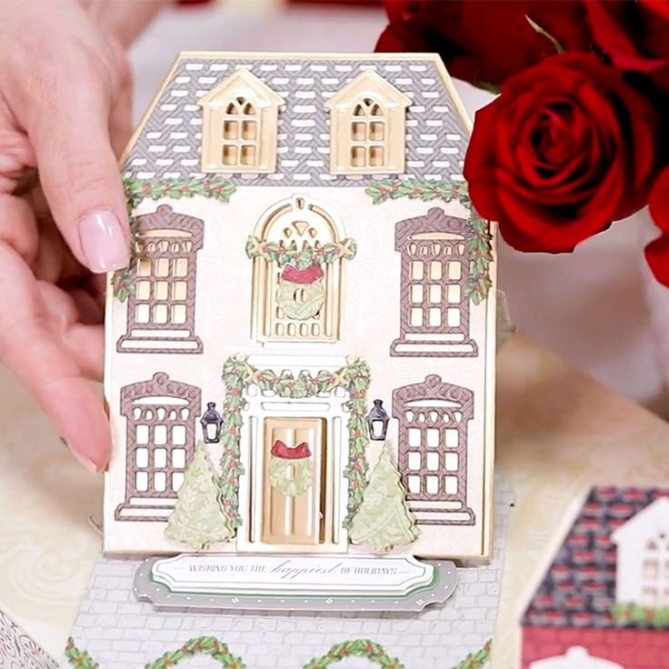 a hand is holding a small doll house with red roses in the foreground and a woman's hands behind it