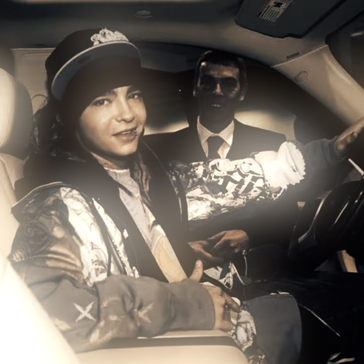 two people sitting in the passenger seat of a car, one is wearing a hat