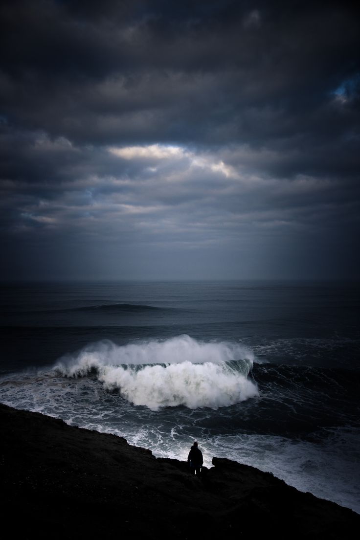 a person sitting on the edge of a cliff looking at an ocean with waves coming in