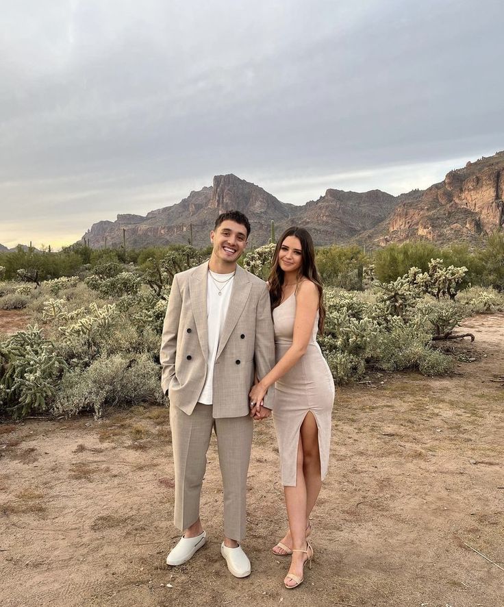 a man and woman standing next to each other in front of mountains with bushes on either side