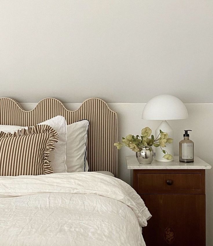 a bed with white linens and striped headboard next to a nightstand with flowers on it