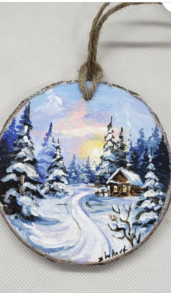 a christmas ornament hanging on a string with snow covered trees and a cabin in the background