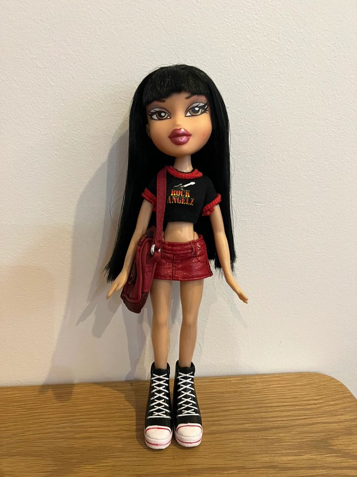 a doll with long black hair wearing a red shirt and skirt on top of a wooden table