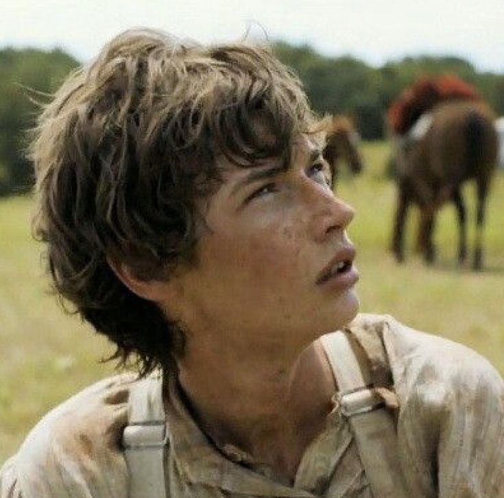 a young man standing in front of horses on a lush green field with trees behind him