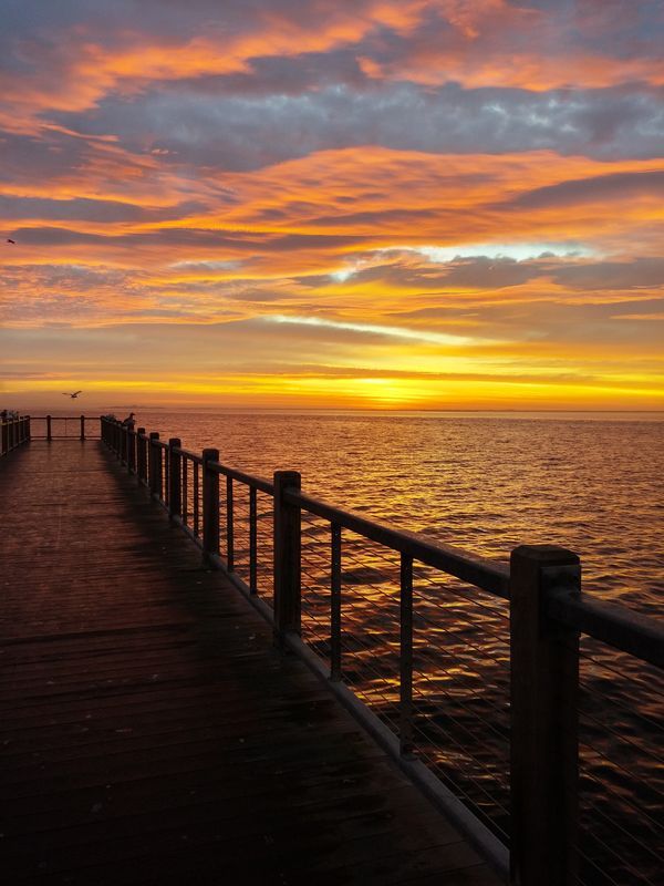 a long pier with the sun setting over the water and clouds in the sky above it