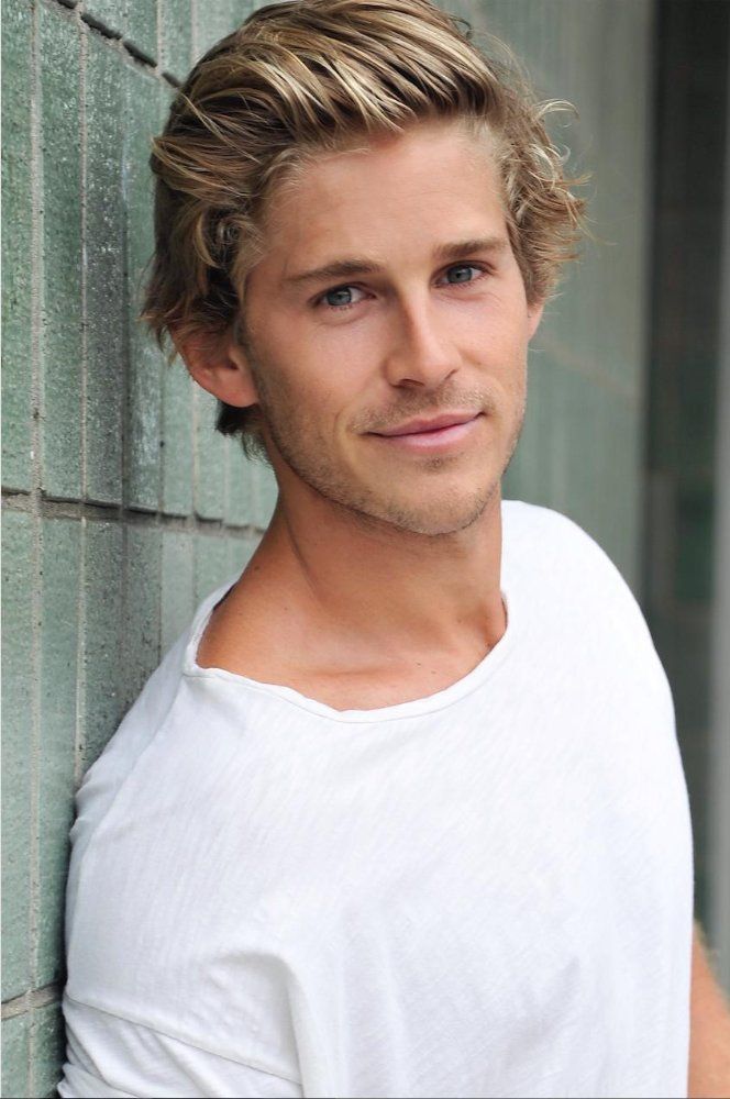 a man leaning against a brick wall wearing a white t - shirt and looking at the camera