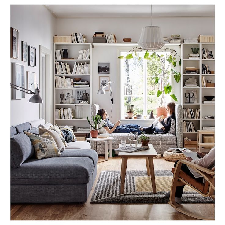 two people sitting in a living room with bookshelves and plants on the wall