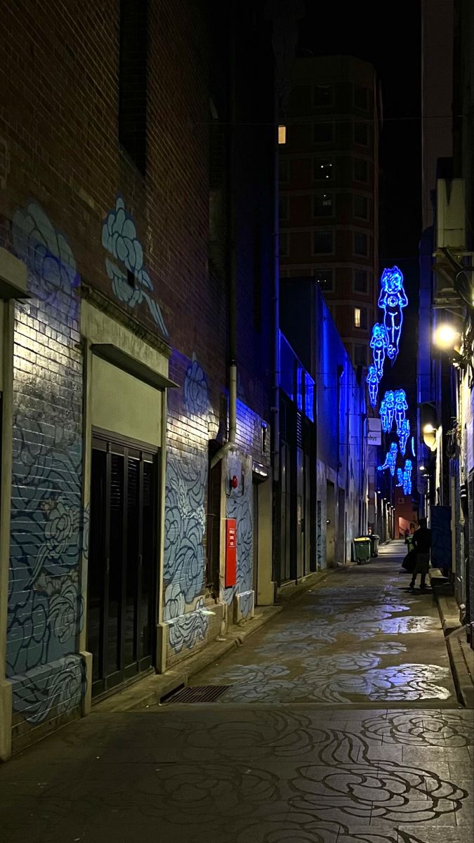 an empty city street at night with graffiti on the buildings and people walking down it