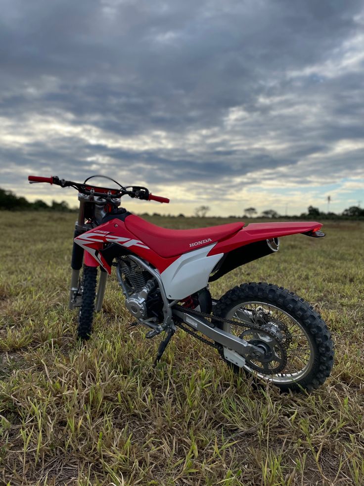 a red and white dirt bike parked in the middle of a field with cloudy skies