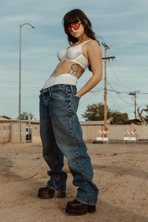 a woman in white shirt and jeans standing on dirt ground with her hands behind her back