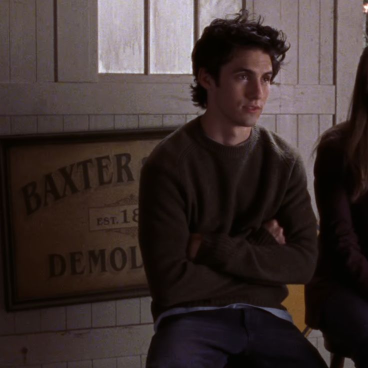 a young man and woman sitting next to each other in front of a wooden sign