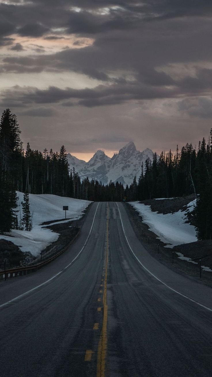 an empty road with snow on the ground and mountains in the background