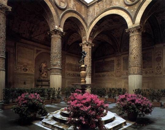 the interior of an old building with pink flowers in pots on the floor and columns