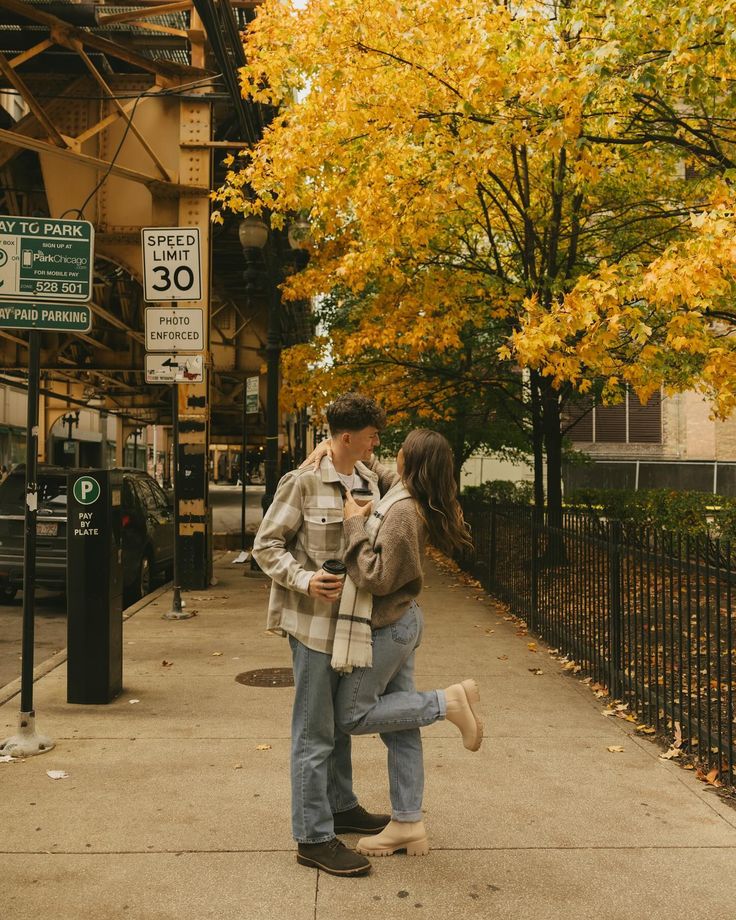 a man and woman are standing on the sidewalk in front of a tree with yellow leaves