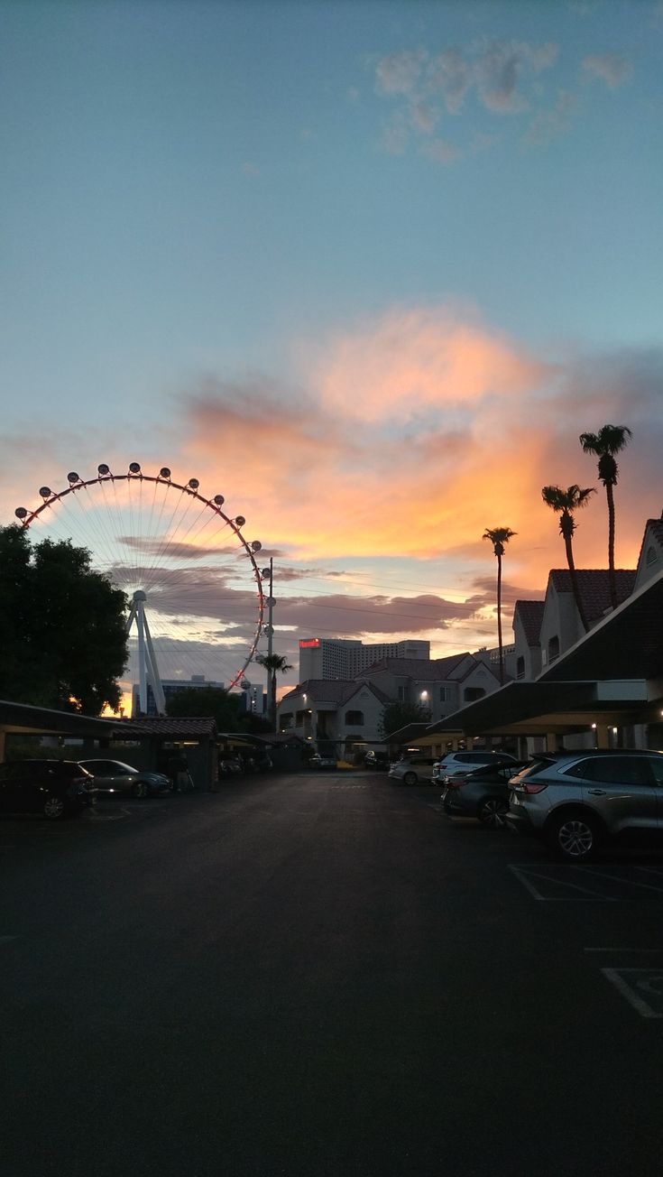 the sun is setting over an amusement park with cars parked on the street and palm trees