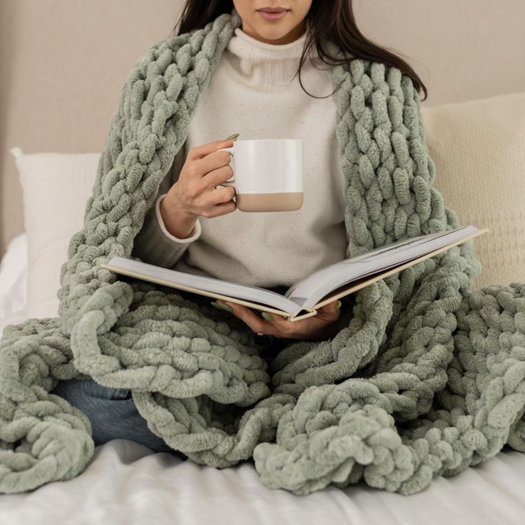 a woman sitting on a bed while holding a coffee cup and reading a book