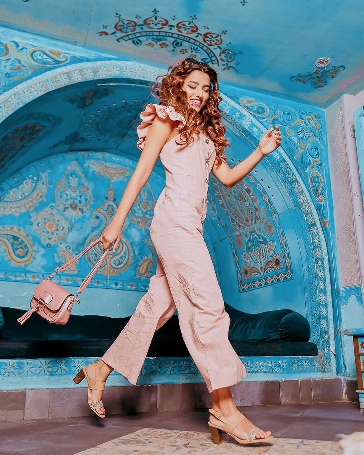 a woman in a pink jumpsuit is holding a handbag and posing for the camera