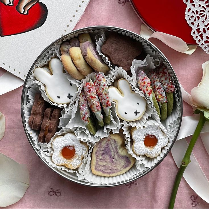 an assortment of cookies and pastries in a tin on a pink table cloth next to flowers