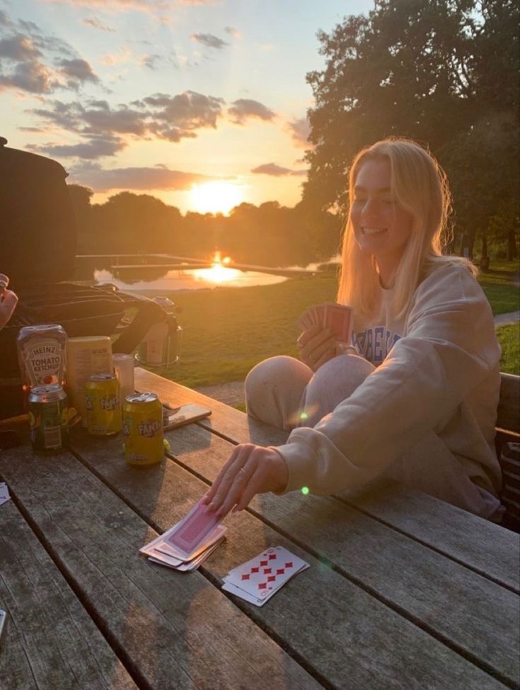 a woman sitting at a picnic table with cards in front of her and the sun setting behind her