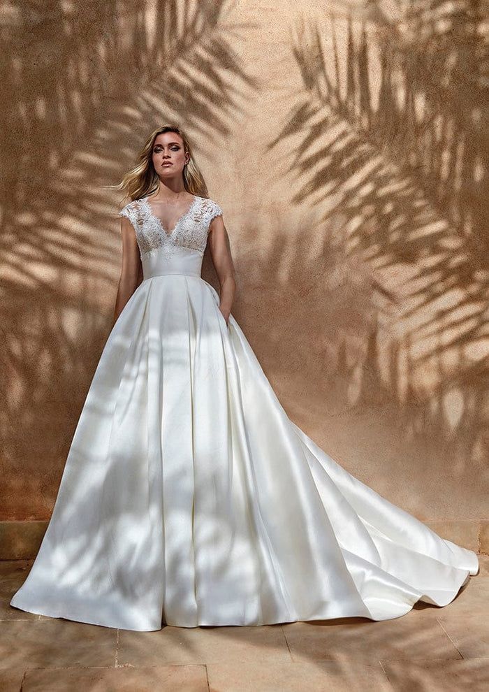 a woman standing in front of a wall wearing a wedding dress with sleeves and pockets