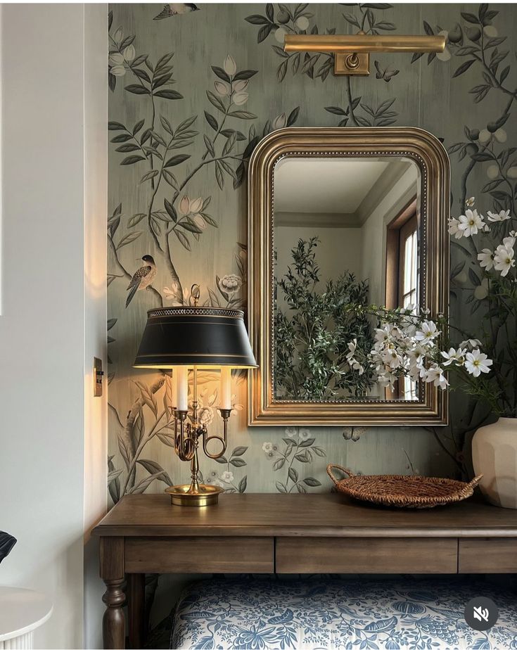 a table with a lamp and mirror on it in front of a floral wallpaper